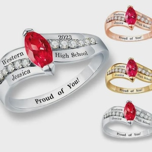 May include: A collection of graduation rings in silver, gold, and rose gold. Each ring features a large, red marquise-cut gemstone and is adorned with small, clear gemstones. The rings are engraved with the words "Western Jessica 2023 High School Proud of You!"
