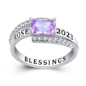 May include: A silver ring with a rectangular, light purple gemstone at the center. The ring is adorned with small, clear stones and the words "ROSE 2023" and "BLESSINGS" are engraved on the band.