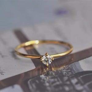 May include: A slender gold ring featuring a small, square-cut diamond. The ring's band is thin and polished, reflecting light. The diamond is set in a four-prong setting. The ring is displayed against a neutral background.