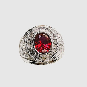 May include: A silver ring with a large, oval, deep red gemstone at its centre. The ring's band features intricate detailing and text around the gemstone. The ring is likely a class ring or commemorative piece.