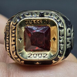 May include: A gold-toned class ring with a large, square, red gemstone at the center. The ring features the text "WASHINGTON UNIVERSITY MEDICINE" and "CLASS OF 2002" in raised lettering. The ring has a black band.