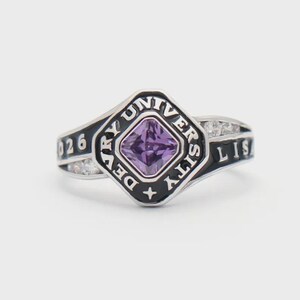 May include: A silver ring featuring a square, purple gemstone. The ring has black enamel detailing with the words "Devry University" and "026" inscribed. Small clear stones are set on the sides.