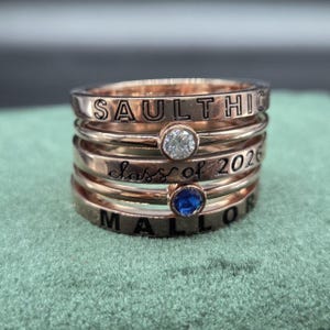 May include: Stack of rose gold rings with engraved text and gemstones. The rings feature the words "SAULT HIGH", "Class of 2026", and "MALLOR". One ring has a clear gemstone, and another has a blue gemstone.