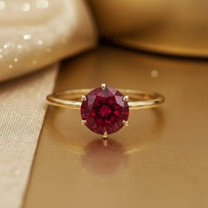 May include: A gold ring with a large, round, deep red gemstone. The gemstone is the focal point, set in a six-prong setting. The ring band is a simple, polished gold. The background is a warm, golden hue.