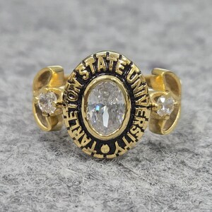 May include: A gold-toned ring featuring a central oval-cut clear stone surrounded by a black border with the text TARLETON STATE UNIVERSITY embossed in gold. Two smaller clear stones are set on the sides of the band, which has a polished finish.