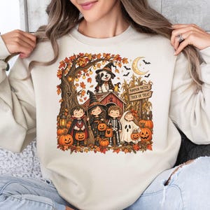 May include: A sand-colored crewneck sweatshirt featuring a colorful Halloween graphic of four children in costumes, a dog in a witch hat, pumpkins, and a wooden sign that reads PUMPKIN PATCH and TRICK OR TREAT, set against a backdrop of autumn trees.
