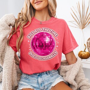 May include: Watermelon-colored t-shirt with a round graphic of a pink disco ball and the text "KISS ALL THE TIME DISCO OCCASIONALLY". The shirt is a short-sleeved, crew neck style.