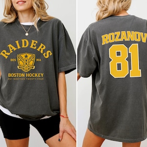 May include: Pepper-colored t-shirt with "RAIDERS" and "BOSTON HOCKEY" text in yellow. The front features a logo with a star, hockey sticks, and the letters "R", "BOS", and "MA". The back displays "ROZANOV 81" in yellow.