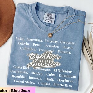 May include: Blue jean Comfort Colors t-shirt with the text "together we are america" and a list of countries. A gold necklace with a pendant is draped across the shirt. The shirt is size S-P-CH.