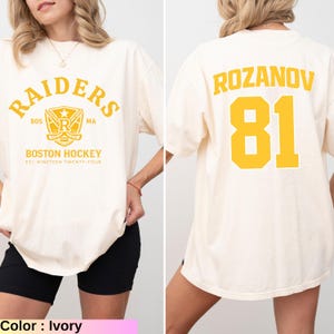 May include: Ivory-colored t-shirt featuring "RAIDERS" arched text above a hockey-themed emblem and "BOSTON HOCKEY" text. The back displays "ROZANOV" and the number "81" in yellow. The shirt's design is a nod to a hockey team.
