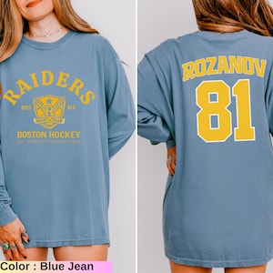 May include: Blue jean long-sleeve shirt with yellow text and graphics. The front features "RAIDERS" and "BOSTON HOCKEY" text. The back displays "ROZANOV" and the number "81" in yellow. The shirt has a crew neck and a relaxed fit.