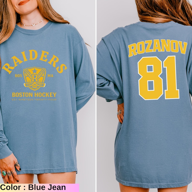 May include: Blue jean long-sleeve shirt with yellow text and graphics. The front features "RAIDERS" and "BOSTON HOCKEY" text. The back displays "ROZANOV" and the number "81" in yellow. The shirt has a crew neck and a relaxed fit.