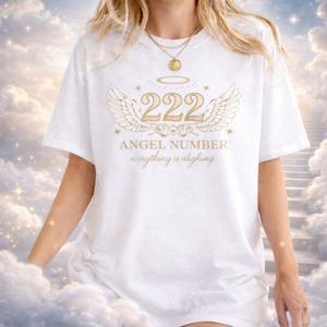 May include: White t-shirt with a gold graphic featuring the angel number 222, wings, and the text "ANGEL NUMBER everything is aligning". The design is centered on the front of the shirt.