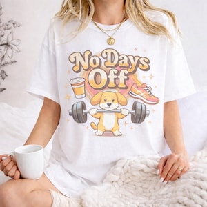 May include: White t-shirt with the text "No Days Off" in a retro font. The graphic features a cartoon dog lifting weights, a coffee cup, and a running shoe. The design is in shades of yellow, orange, and brown.