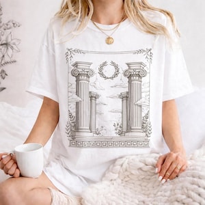 May include: White t-shirt featuring a black and white illustration of classical columns, clouds, and a laurel wreath. The design is centered on the shirt, with decorative borders. The shirt is a casual, oversized fit.