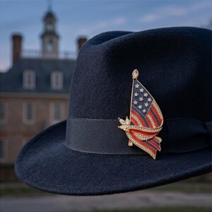 Op de afbeelding: Een donkerblauwe vilten fedora-hoed met een brede grosgrain lintband. Op de band is een goudkleurige metalen broche bevestigd in de vorm van een wapperende Amerikaanse vlag, versierd met rode en blauwe emaille strepen en heldere strass-steentjes.