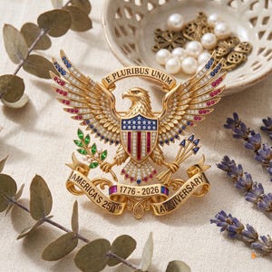 Pu&ograve; includere: Una spilla dorata a forma di aquila con il testo E PLURIBUS UNUM sopra la testa e AMERICA'S 250TH ANNIVERSARY 1776-2026 su un nastro sottostante. L'aquila &egrave; decorata con pietre rosse, bianche e blu, foglie verdi e cristalli trasparenti.