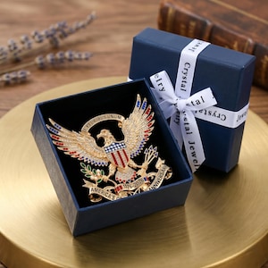 Pu&ograve; includere: Una spilla a forma di aquila color oro con smalto rosso, bianco e blu e cristalli trasparenti. L'aquila tiene frecce e un ramoscello d'ulivo, con un nastro che reca la scritta E PLURIBUS UNUM e AMERICA'S 250TH ANNIVERSARY 1776-2026, in una scatola blu.