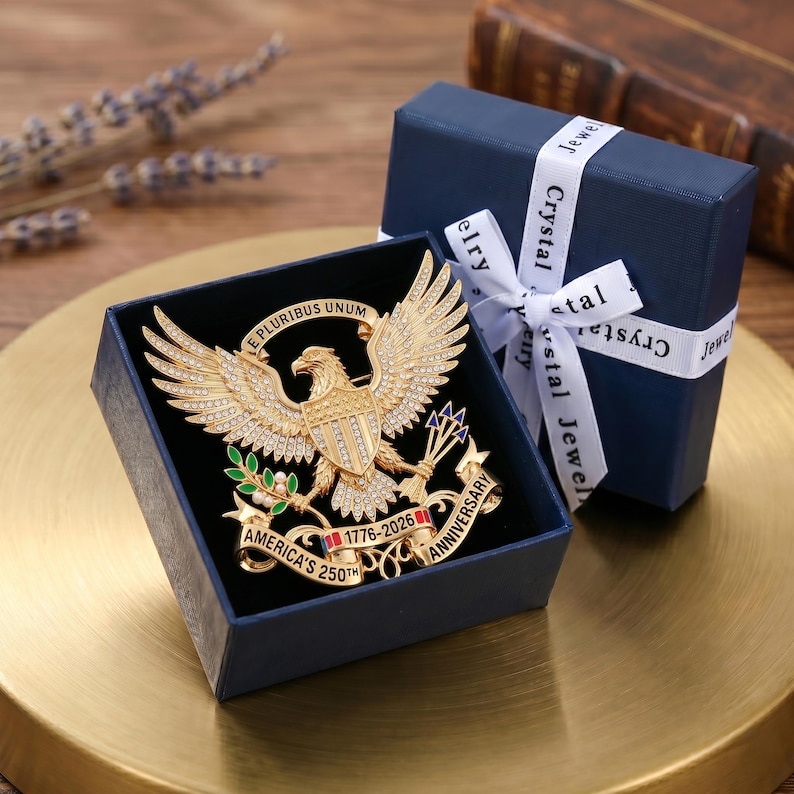 Pu&ograve; includere: Una spilla a forma di aquila color oro con cristalli trasparenti, uno scudo sul petto, foglie verdi e un nastro con la scritta E PLURIBUS UNUM e AMERICA'S 250TH ANNIVERSARY 1776-2026. La spilla &egrave; contenuta in una scatola regalo blu scuro.