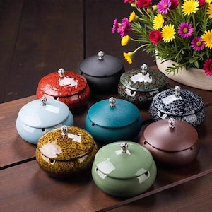 Puede incluir: Colección de ceniceros de cerámica en varios colores, incluyendo rojo, azul, verde y marrón. Cada cenicero tiene una tapa con un pomo decorativo. Los ceniceros están dispuestos sobre una superficie de madera, con un jarrón de flores coloridas en el fondo.