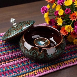 Puede incluir: Un cenicero de cerámica con tapa, base marrón con esmalte moteado en verde azulado y marrón. La tapa está parcialmente abierta, revelando el interior y un pequeño cenicero central. Un pomo decorativo plateado adorna la tapa. Un colorido ramo de flores en el fondo.