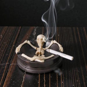 Op de afbeelding: Een novelty asbak met een skeletfiguur die in een donkerbruine asbak ligt. Een brandende sigaret rust in de asbak, met opstijgende rook. De asbak staat op een donker houten oppervlak.