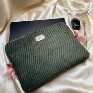 Peut inclure: Une housse pour ordinateur portable en velours côtelé vert olive foncé avec une étiquette en tissu blanc rectangulaire portant le texte LFT imprimé en noir. La housse est ornée de rubans en satin rose clair aux coins, une souris noire et un stylo doré.