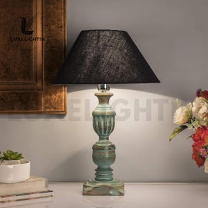 May include: A teal and black table lamp with a distressed finish. The lamp features a black lampshade and a base with an urn shape. The lamp is next to a bouquet of flowers and a small succulent plant.