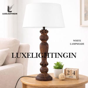 Puede incluir: Una lámpara de mesa de madera con una pantalla blanca. La lámpara tiene una base de madera tallada de color marrón oscuro y una pantalla blanca. El texto "LUXELIGHTINGIN" se muestra en la lámpara. Una pequeña planta, una bola decorativa y un calendario de madera están sobre la mesa.