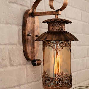 Puede incluir: Aplique de pared de estilo antiguo con acabado bronce. La lámpara presenta un diseño ornamental con un cilindro de vidrio que alberga una bombilla de estilo vintage. El aplique está montado en una pared de ladrillo.