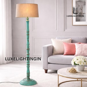 Puede incluir: Una lámpara de pie con una base turquesa envejecida y una pantalla beige. La lámpara tiene una base redonda y una columna decorativa. La pantalla es de color neutro. La lámpara está colocada sobre una alfombra de color claro. La altura de la lámpara es de unos 150 cm.