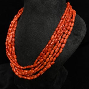 May include: A multi-strand necklace composed of vibrant red coral beads. The beads are barrel-shaped and strung together, creating a layered effect. The necklace is displayed on a black bust, showcasing the jewelry's color and texture.