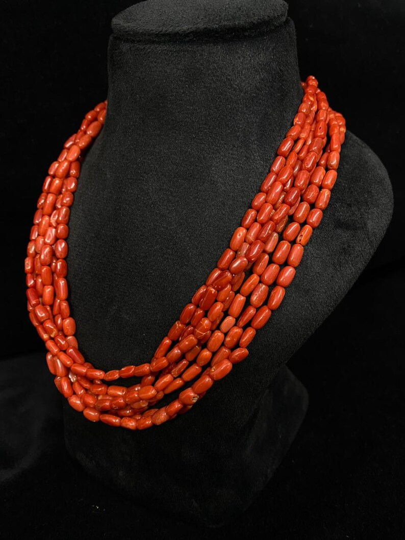 May include: A multi-strand necklace composed of vibrant red coral beads. The beads are barrel-shaped and strung together, creating a layered effect. The necklace is displayed on a black bust, showcasing the jewelry's color and texture.