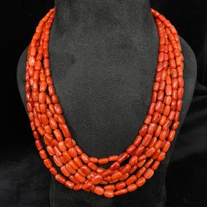 May include: A multi-strand necklace composed of vibrant, reddish-orange coral beads. The necklace is displayed on a black bust, showcasing the layered design and natural texture of the coral.