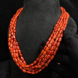 May include: A multi-strand necklace featuring vibrant orange-red coral beads. The necklace is displayed on a black mannequin, showcasing the color and texture of the polished coral. The beads are oval-shaped and strung together.
