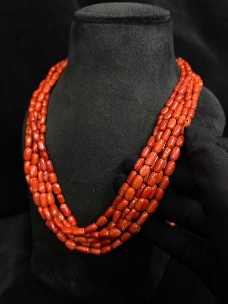 May include: A multi-strand necklace featuring vibrant orange-red coral beads. The necklace is displayed on a black mannequin, showcasing the color and texture of the polished coral. The beads are oval-shaped and strung together.