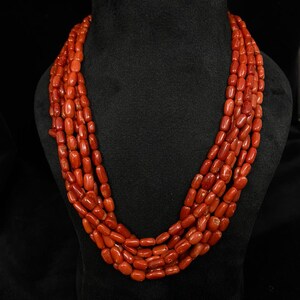 May include: A multi-strand necklace composed of vibrant red coral beads. The beads are oval-shaped and strung together, creating a layered effect. The necklace is displayed on a black mannequin, highlighting the color and texture of the coral.