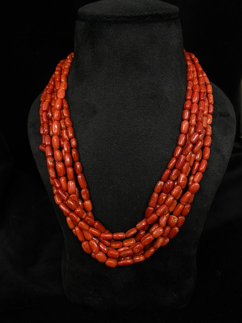 May include: A multi-strand necklace composed of vibrant red coral beads. The beads are oval-shaped and strung together, creating a layered effect. The necklace is displayed on a black mannequin, highlighting the color and texture of the coral.