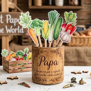 Può includere: Un portapenne in legno con la scritta "Papa" e un design a tema vegetale. Il portapenne contiene ritagli di verdure su bastoncini, tra cui carote, broccoli, mais e altro. È presente anche una cassa di legno con ritagli di verdure.