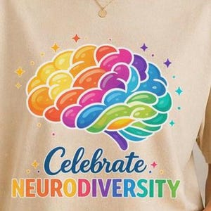 May include: A beige t-shirt featuring a colorful brain graphic with the words "Celebrate Neurodiversity." The brain illustration uses a rainbow of colors, including yellow, orange, pink, purple, and blue. The text is in a playful, multi-colored font.