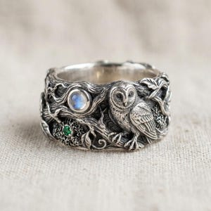 May include: A wide silver ring featuring a detailed relief of an owl perched on tree branches. The band includes a round, iridescent moonstone cabochon and a small, circular green gemstone set among the textured, oxidised silver metalwork.