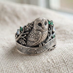 May include: A silver ring featuring a detailed owl perched on a branch, surrounded by oak leaves and small green and iridescent stones. The band is designed as a musical staff with a treble clef and small clear stones embedded along the lines.