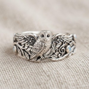 Peut inclure: Une bague en argent représentant un hibou détaillé perché parmi des feuilles sculptées et des notes de musique. L'anneau comprend de petites pierres rondes et irisées incrustées dans le métal, illustrant un hibou texturé aux yeux sombres.