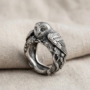 May include: A silver ring featuring a detailed owl perched atop a band sculpted to resemble intertwined oak branches with textured leaves and small acorns. The metal has an oxidised finish that highlights the intricate carvings of the bird.