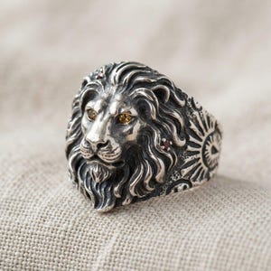 May include: A sterling silver ring featuring a detailed lion head with a flowing mane. The lion has two amber-colored eyes and small red gemstones embedded within the textured silver mane. The band displays intricate sunburst patterns on the sides.