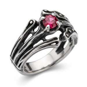 Peut inclure: Une bague en argent au design complexe et organique. Elle présente une pierre précieuse rouge vif et ronde en son centre. Le travail du métal a une apparence texturée, presque squelettique, avec des lignes fluides et des espaces ouverts. La bague est sur fond blanc.