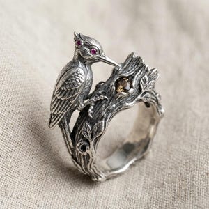 Könnte beinhalten: Ein silberner Ring in Form eines strukturierten Baumastes mit einem detaillierten Vogel an der Seite. Der Vogel hat kleine rosa Edelsteinaugen und der Ast ist mit einem einzelnen runden braunen Edelstein verziert, der in die Rinde eingelassen ist.