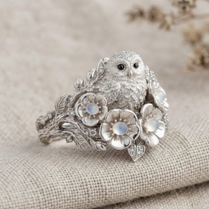 May include: A silver ring featuring a detailed owl figure perched among sculpted branches and leaves. The design includes three prominent flowers with iridescent moonstone centres and small clear stones embedded throughout the metal surface.
