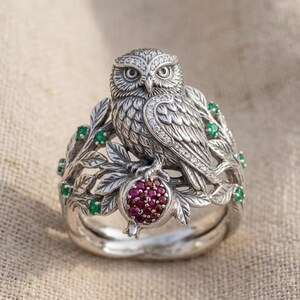 May include: A silver ring featuring a detailed owl perched on a branch with leaves. The owl is accented with small white stones along its wing. The branch includes small green stones and a central pomegranate fruit set with multiple red stones.