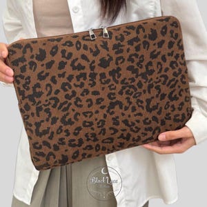 May include: Brown laptop sleeve with a corduroy texture and a black leopard print pattern. The sleeve has a zipper closure and is designed to protect a laptop. The overall aesthetic is stylish and modern.
